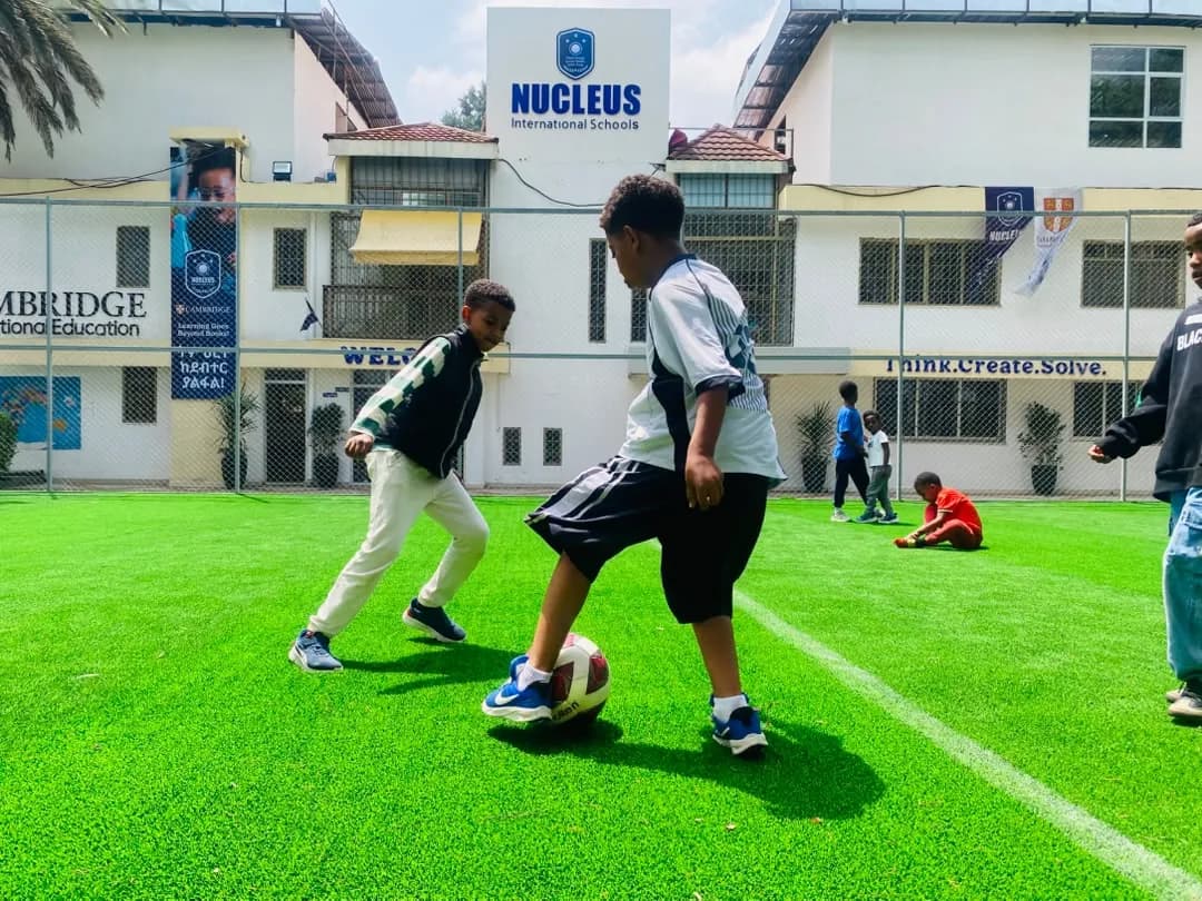 Two campers contesting a football on the artificial turf in front of the Nucleus International Schools building