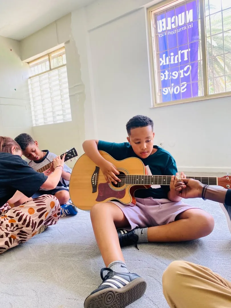A camper concentrating on an acoustic guitar chord beneath a Nucleus banner in the music room