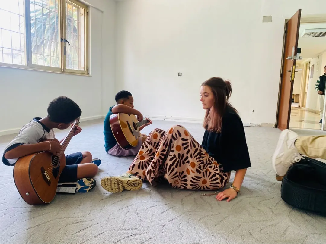 A music teacher sitting on the floor guiding two campers holding acoustic guitars