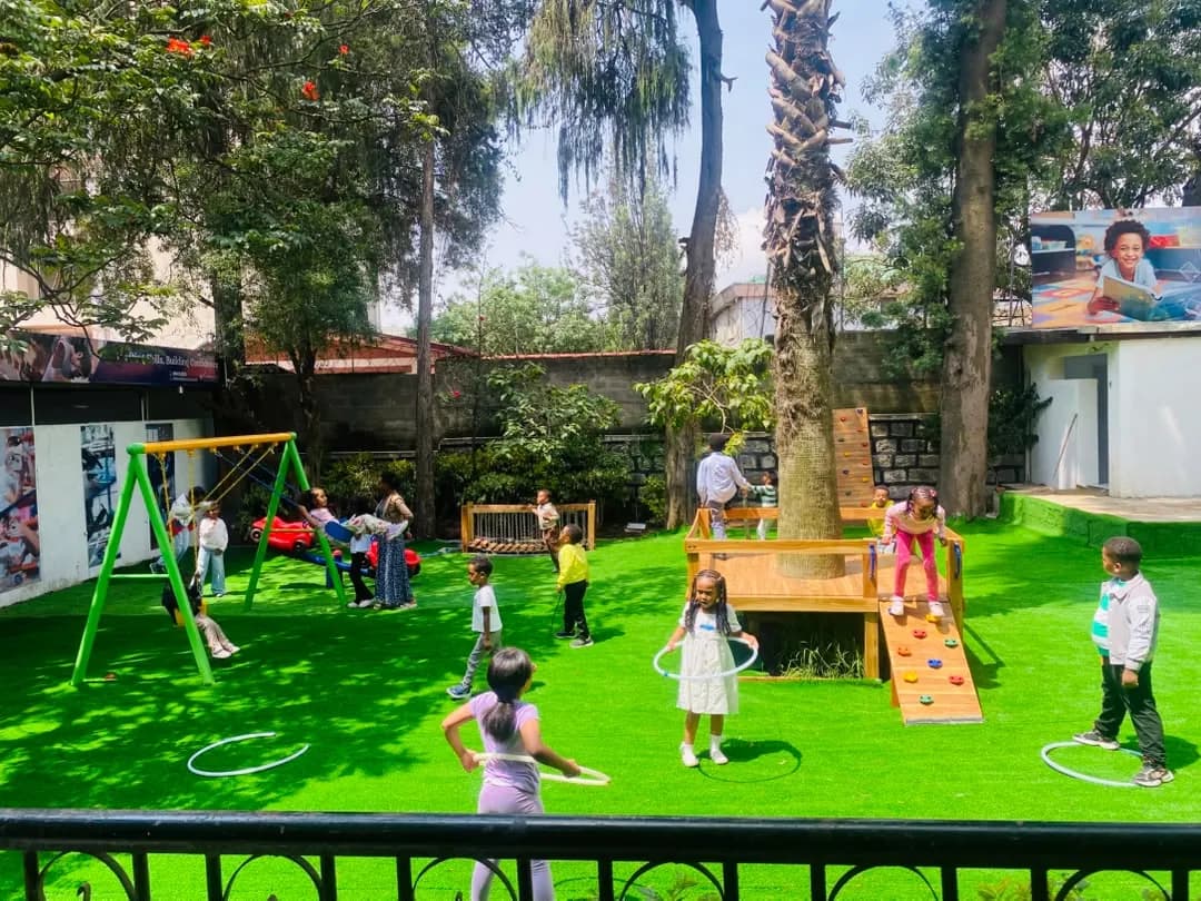 Campers playing with hula hoops on the green playground under the big tree, with swings and a climbing frame behind them