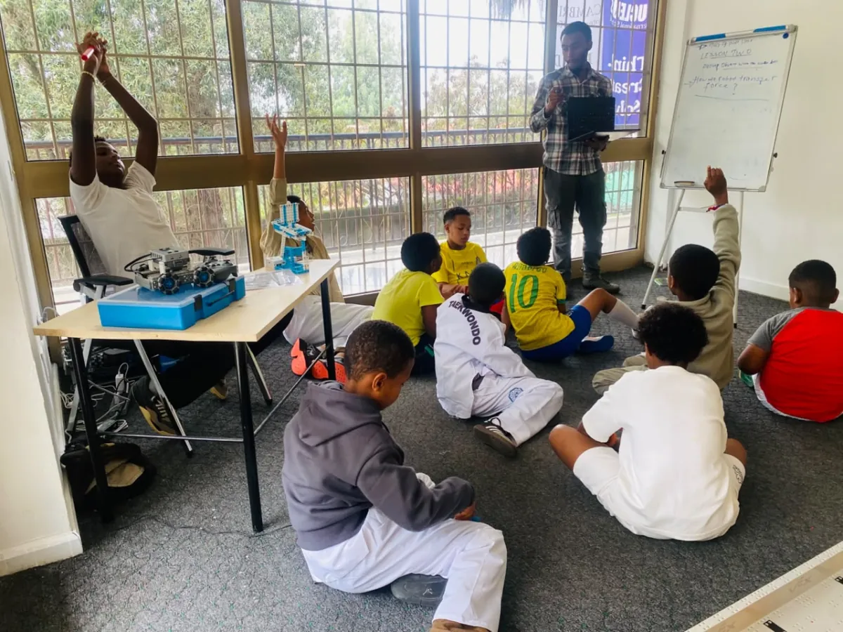 A robotics instructor holding up a robot part while campers raise their hands during robotics class