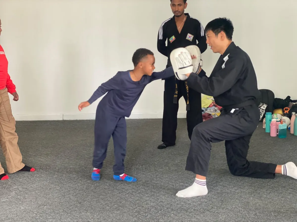 A young camper striking a training pad held by the taekwondo instructor while a coach watches
