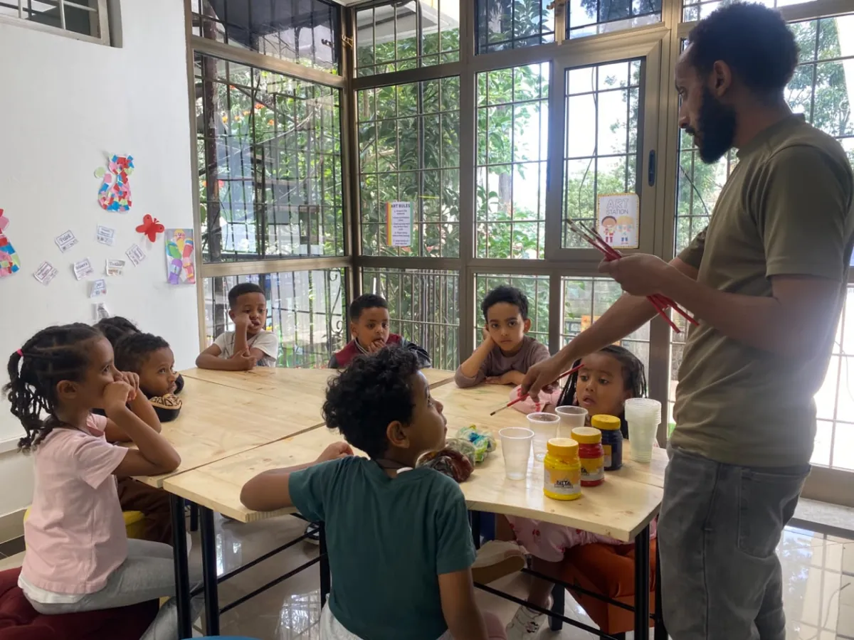 The art teacher holding up two brushes while campers wait at the paint table by the window