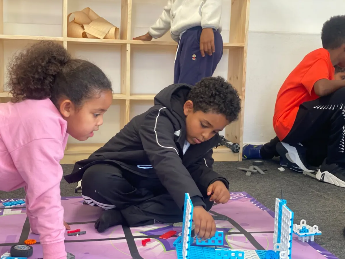 Two campers assembling a blue tower on the printed city mat while classmates build behind them
