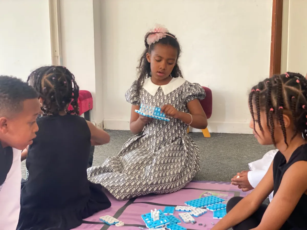 A camper holding up a strip of coding tiles and explaining it to the friends sitting around her on the activity mat