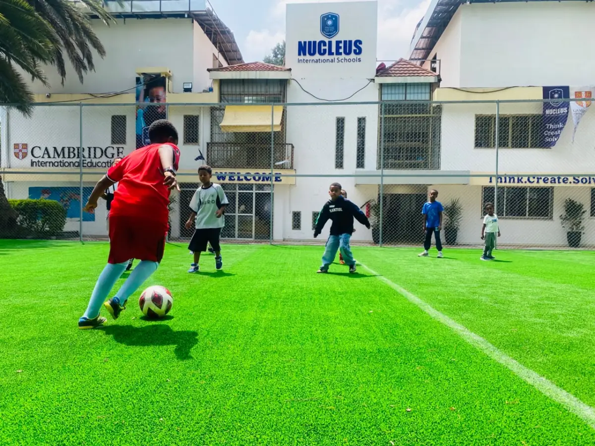 A camper in red driving the ball forward on the turf in front of the school building