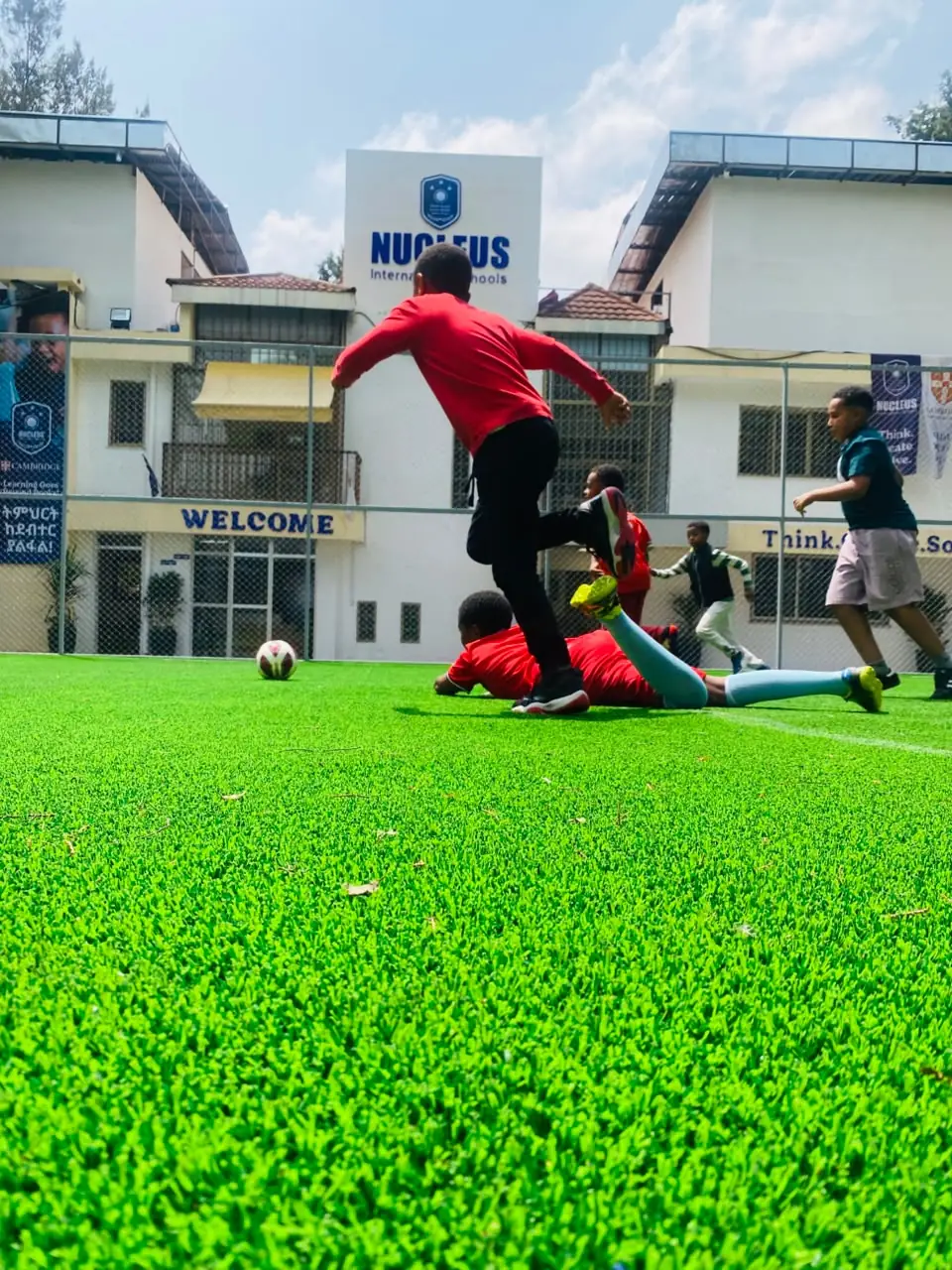 Two campers going to ground for the ball on the turf in front of the Nucleus International Schools building