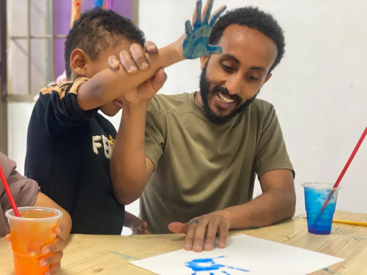 A teacher laughing with a camper as he presses his paint covered hand onto paper