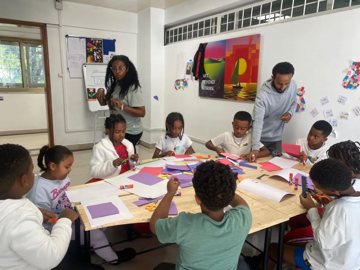 Two art teachers guiding a full table of campers cutting and gluing coloured paper shapes