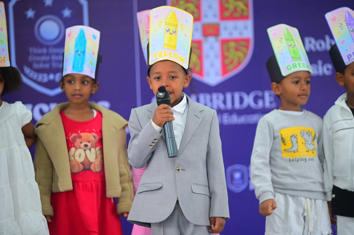 A young camper in a grey suit singing into a microphone on stage while the rest of his group stands behind him in paper crayon hats