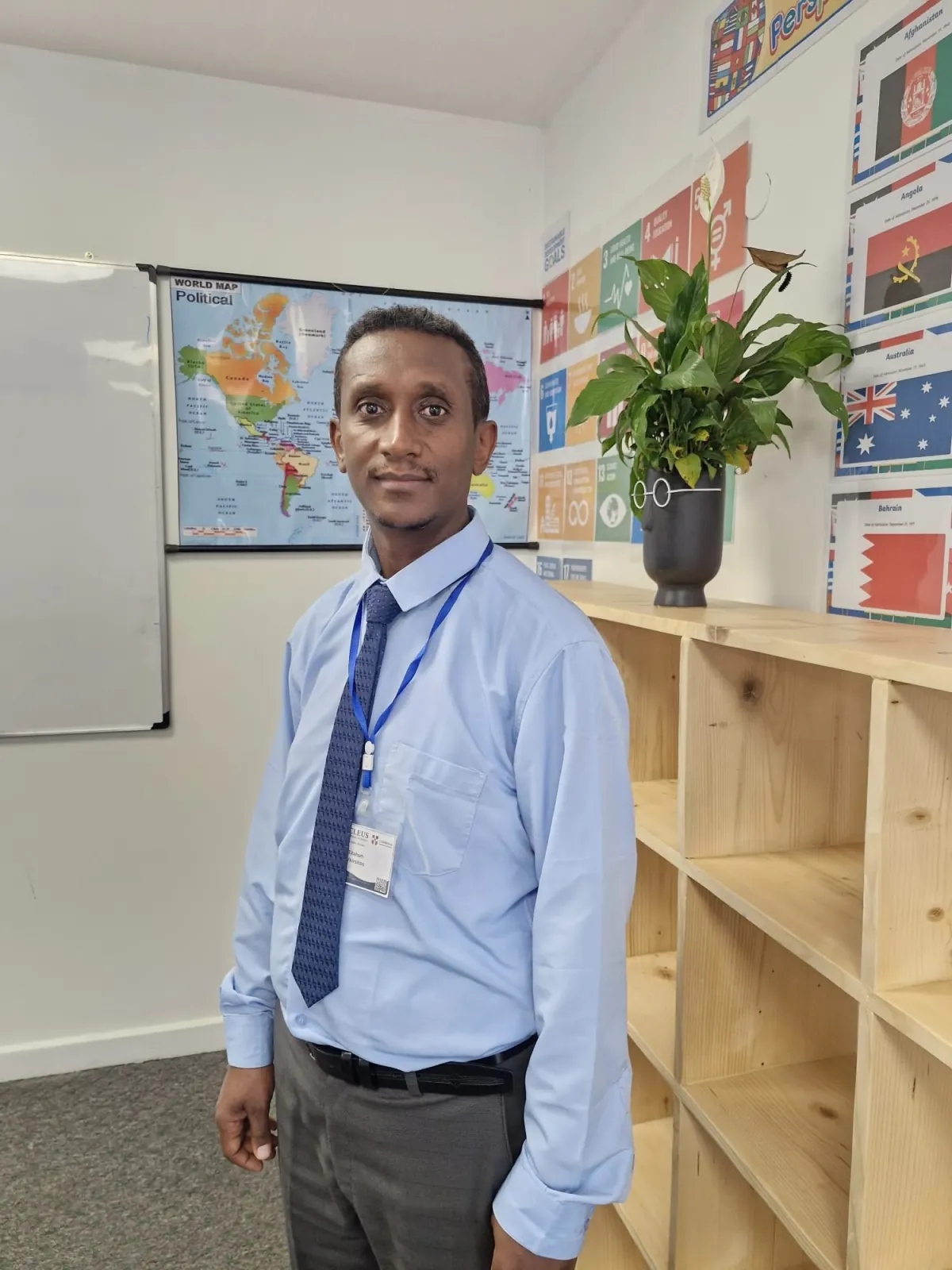 Tilahun G/Kiristose standing in the Global Perspectives classroom at Nucleus International School, in front of a world map and a wall of national flags