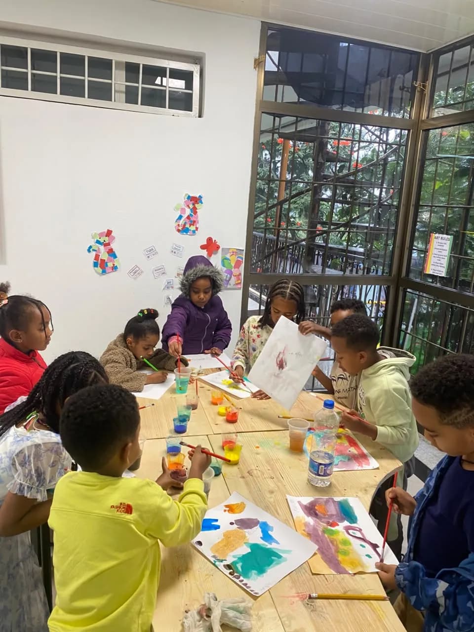 Campers standing around the wooden art table painting, one holding up a finished sheet to show the group