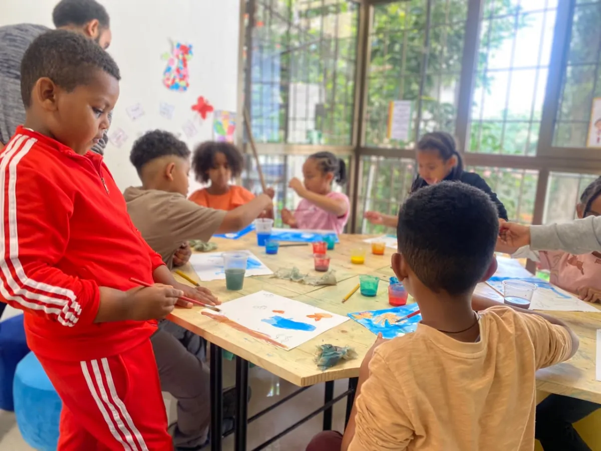 A camper in a red tracksuit painting at the art table while a teacher helps the children around him