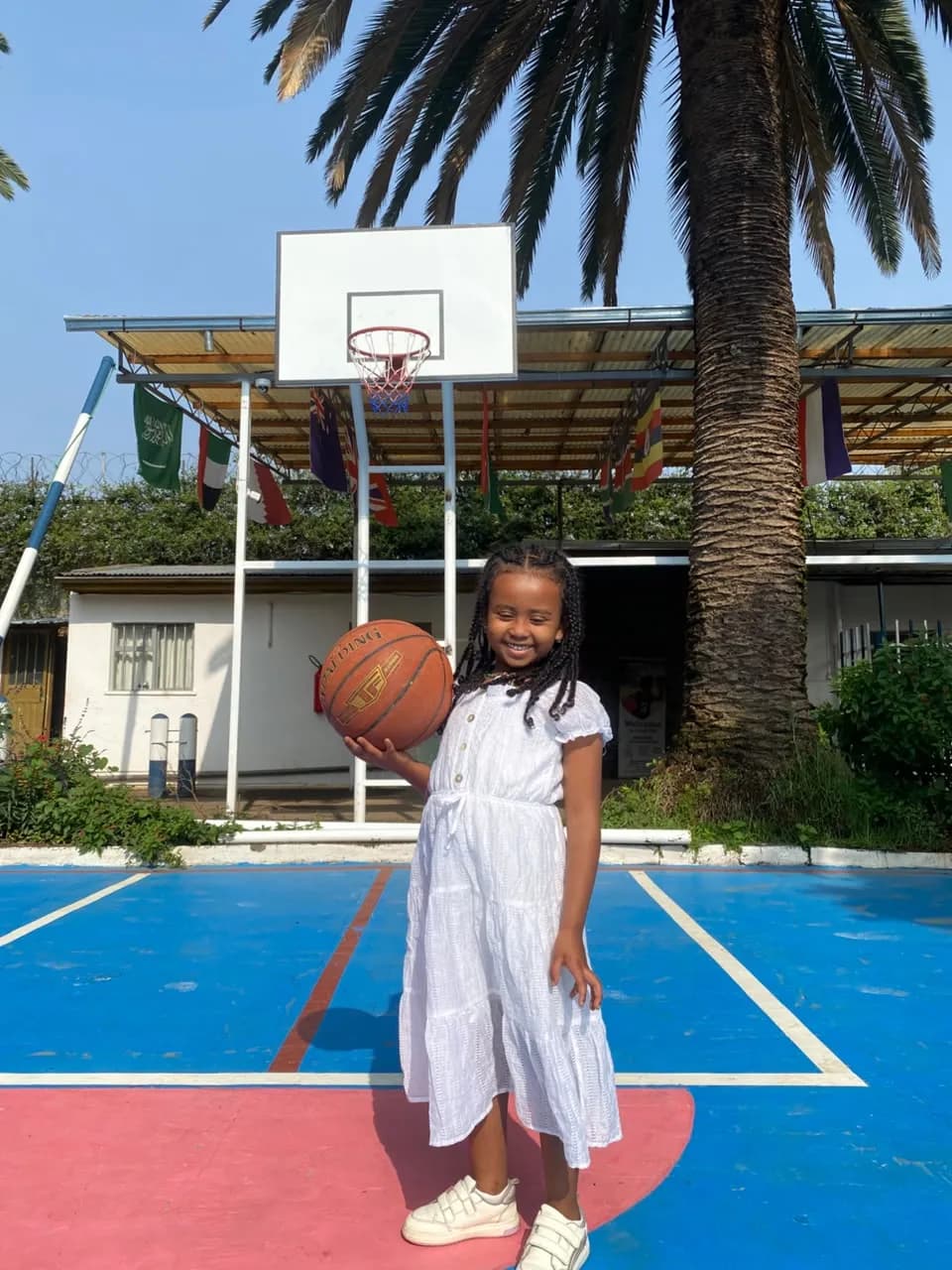 A young camper in a white dress standing on the blue basketball court holding a basketball and smiling