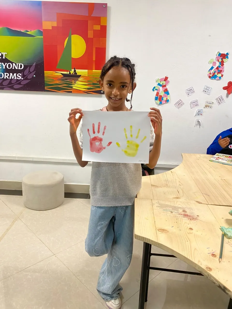 A camper holding up her handprint painting in front of the Art Beyond Norms poster in the art room