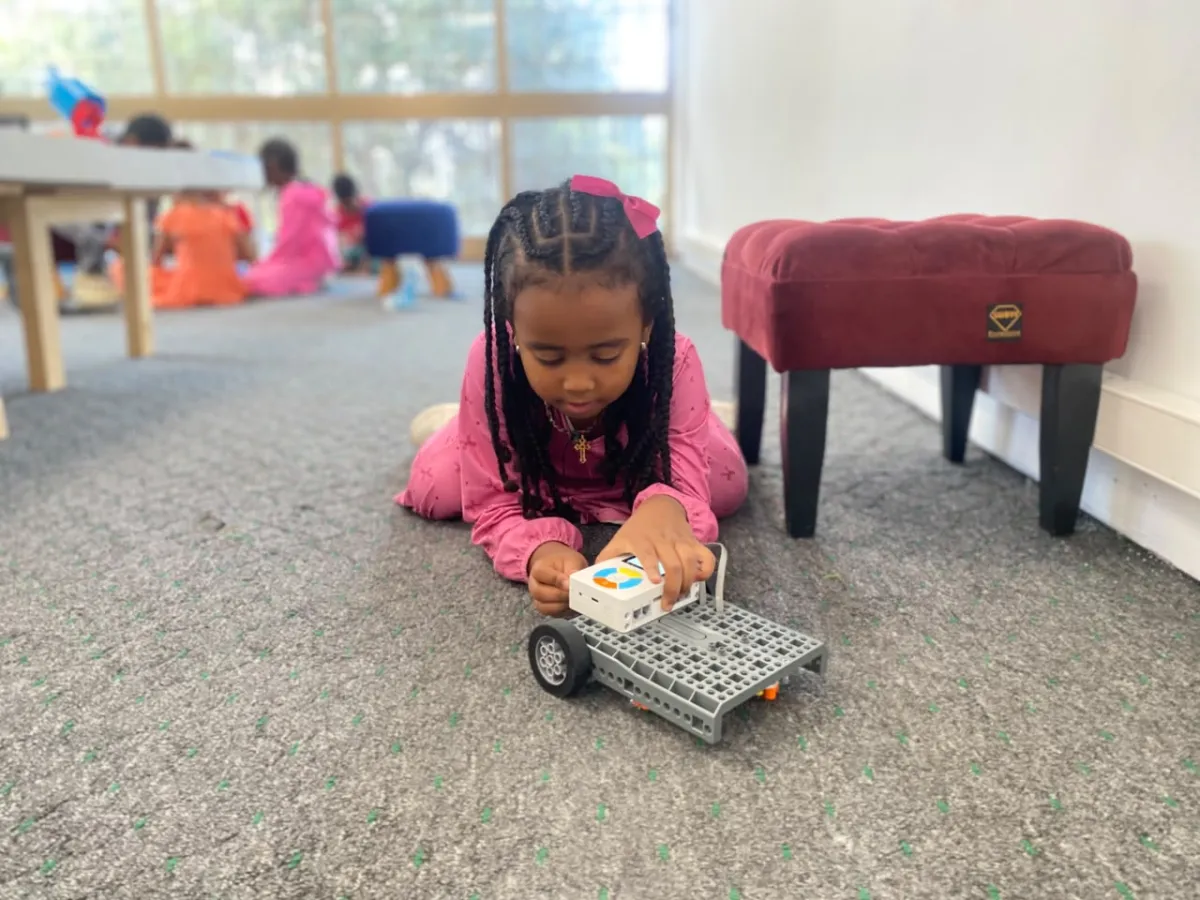A young camper lying on the carpet driving a small wheeled robot she has just finished building