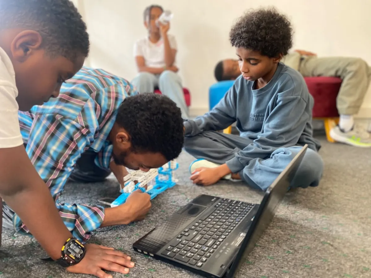 An instructor kneeling on the floor helping two campers assemble a robotics kit next to an open laptop